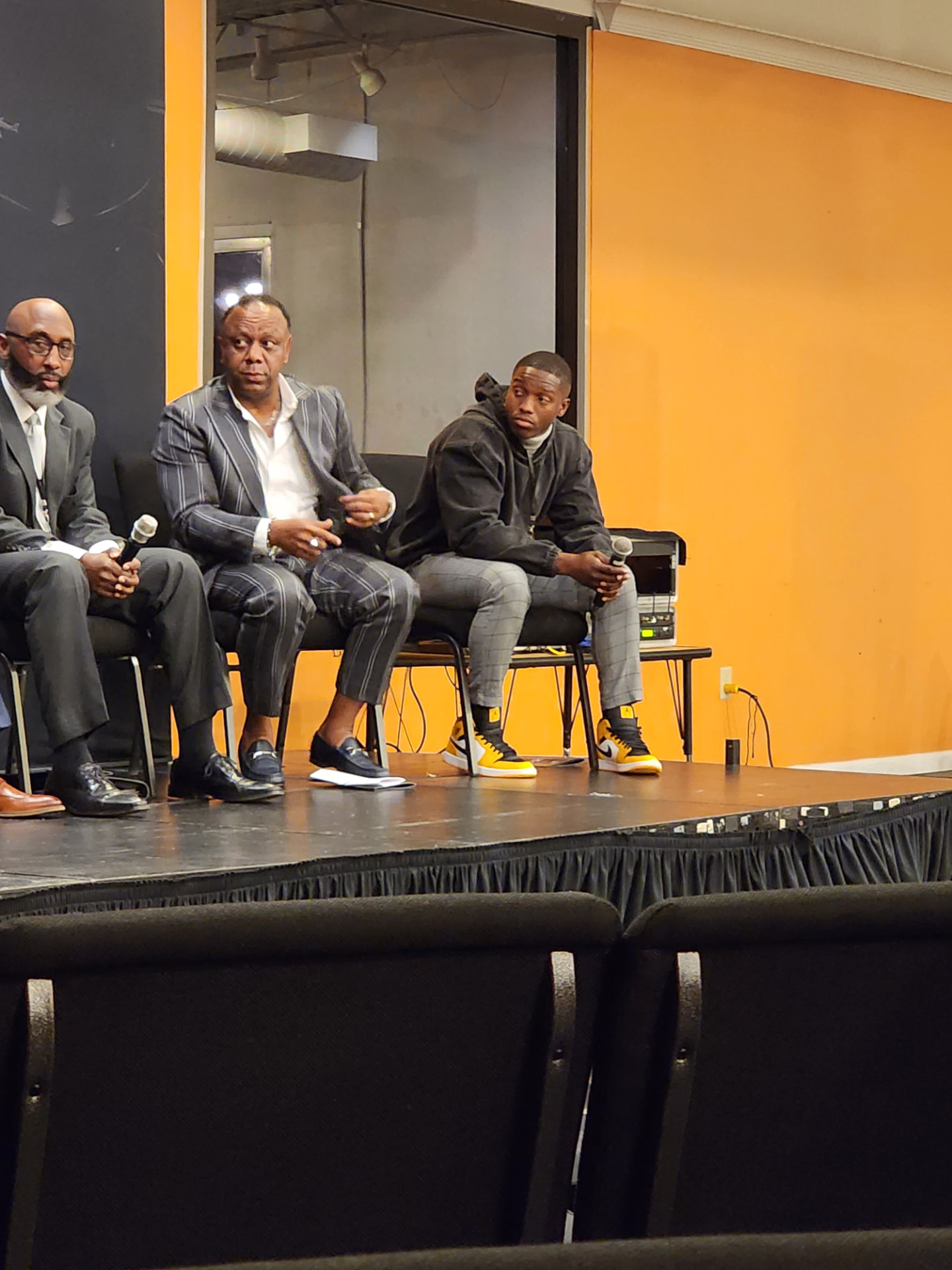 A three-man panel on stage, including a young adult panelist, holding microphones